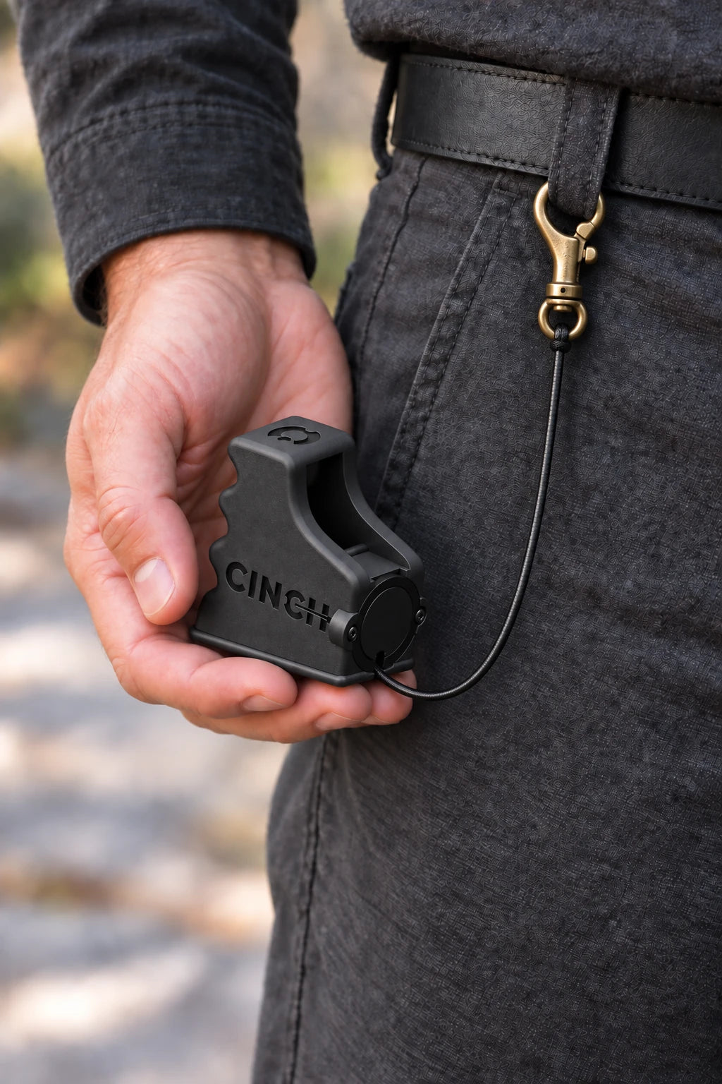 Belt carry: A hand holds the matte black Cinch V15 at the hip, with its bottom-exit cord attached to a brass clip on a belt loop. Product visualization.