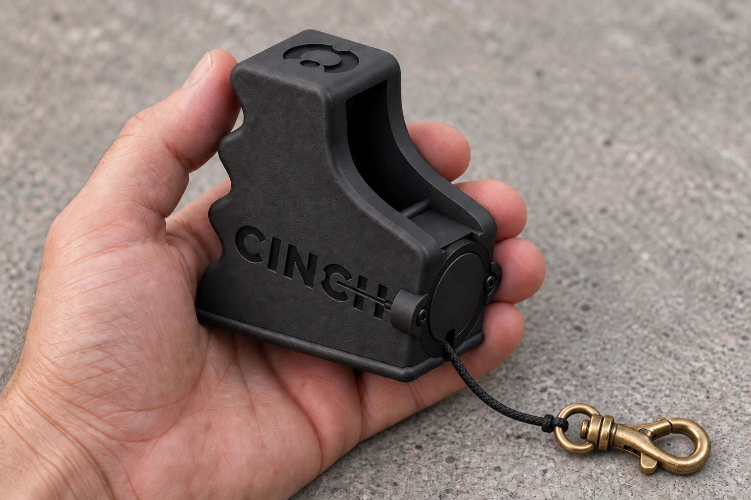 In the hand: A matte black Cinch V15 rests in one hand, with its cord exiting below the reel and a brass swivel clip. Product visualization.