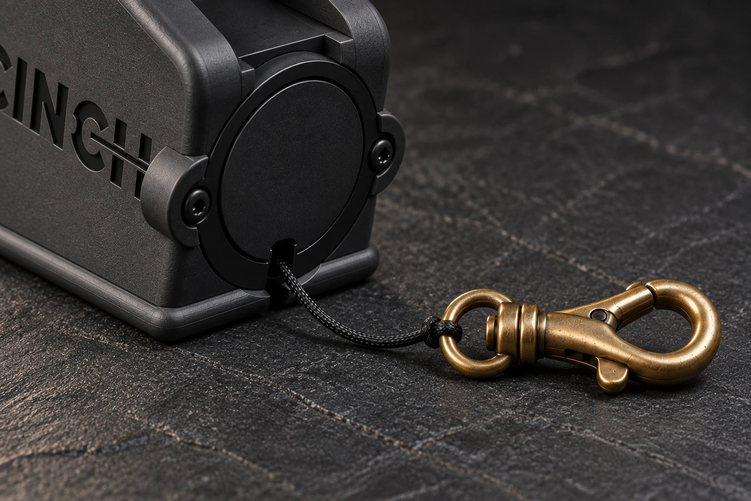 Reel and clip: Close view of the seated black reel, two mounting screws, bottom-exit cord and brass swivel clip. Product visualization.